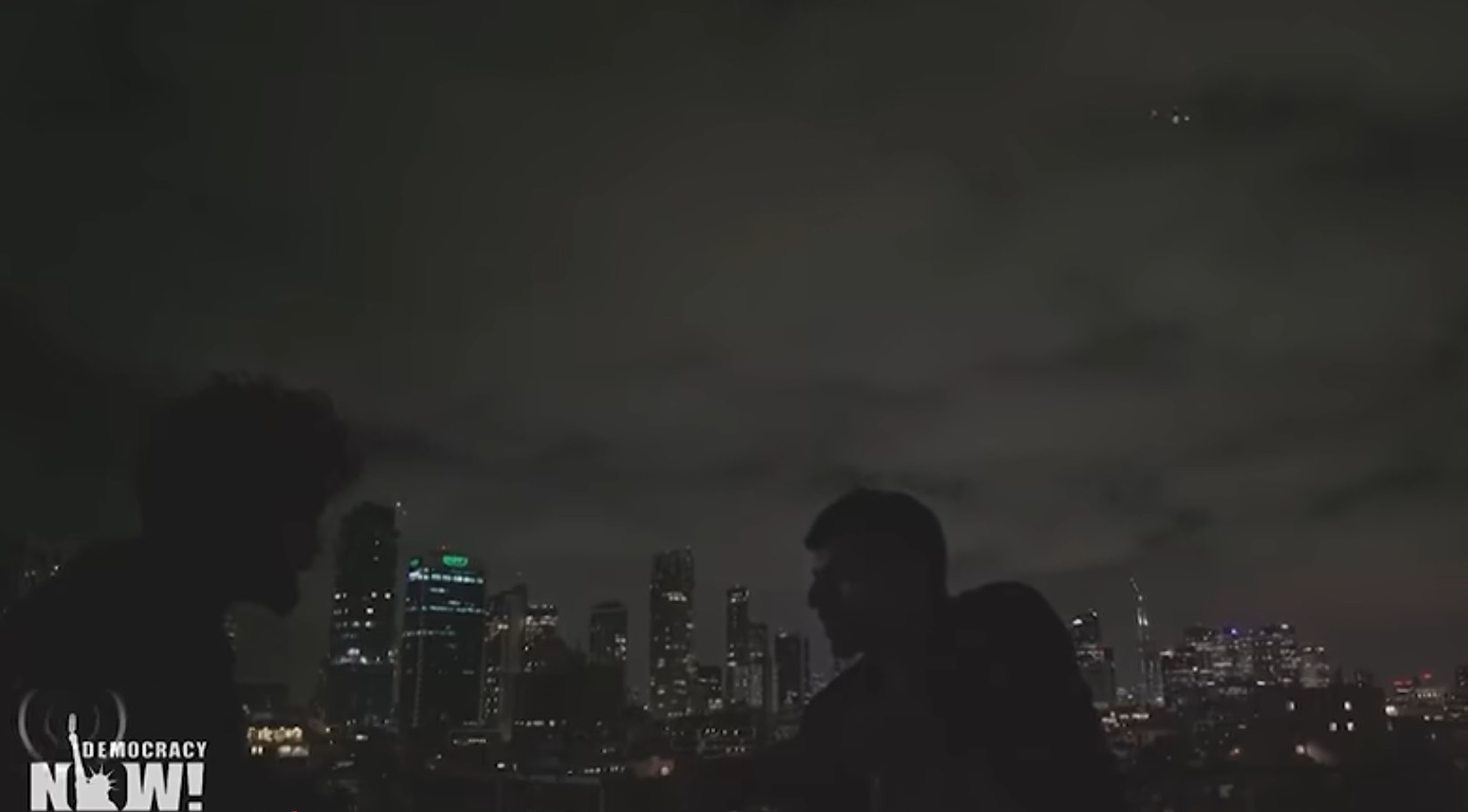 Standbild aus dem Film: zwei anonymisierte Gestalten auf einer Dachterrasse vor der Skyline von Tel Aviv bei Nacht